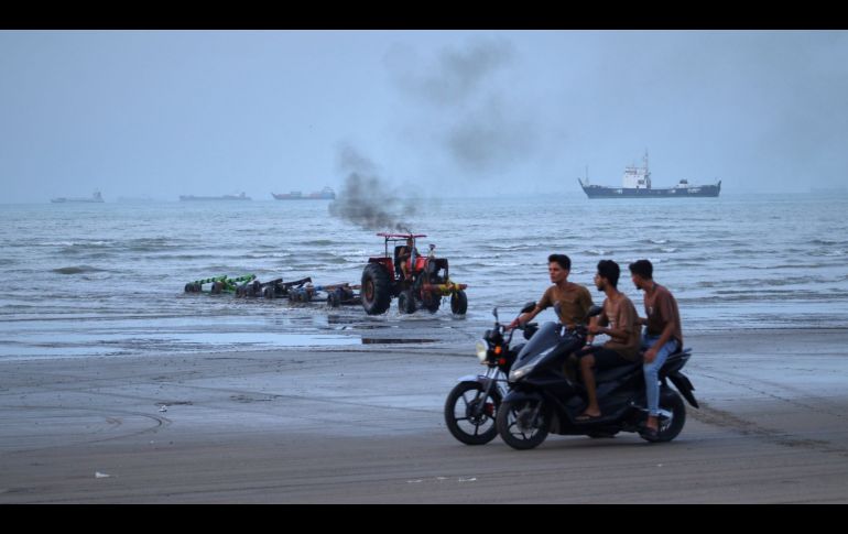 Jóvenes a bordo de motocicletas a lo largo de la orilla mientras un tractor saca equipo del agua y al fondo se pueden ver buques mercantes aparentemente anclados en el estrecho de Ormuz. AP/R. Poudat